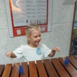 Dziewczynka stoi przy ksylofonie drewnianym i próbuje na nim grać pałeczkami