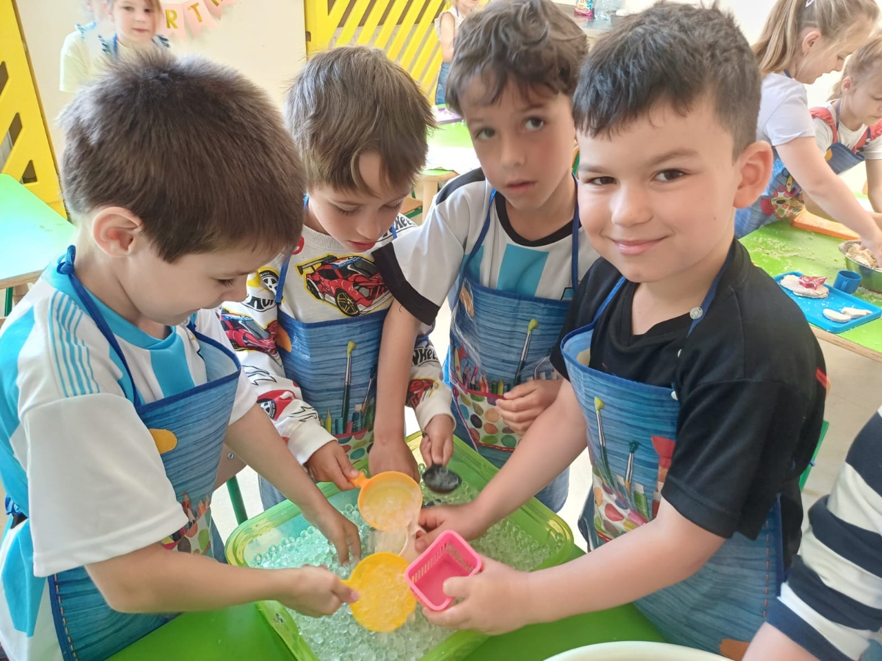 Zabawy sensoryczne - Dzieci przy stole bawią się małymi, szklanymi kuleczkami umieszczonymi w zielonym pojemniku na stole