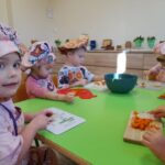 Zdjęcie przedstawia dzieci siedzące przy stoliku i krojące składniki na śląski szałot.